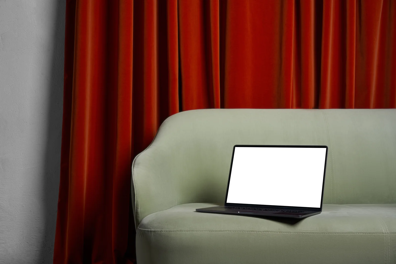 Portátil con la pantalla en blanco sobre un sofá verde junto a unas cortinas rojas