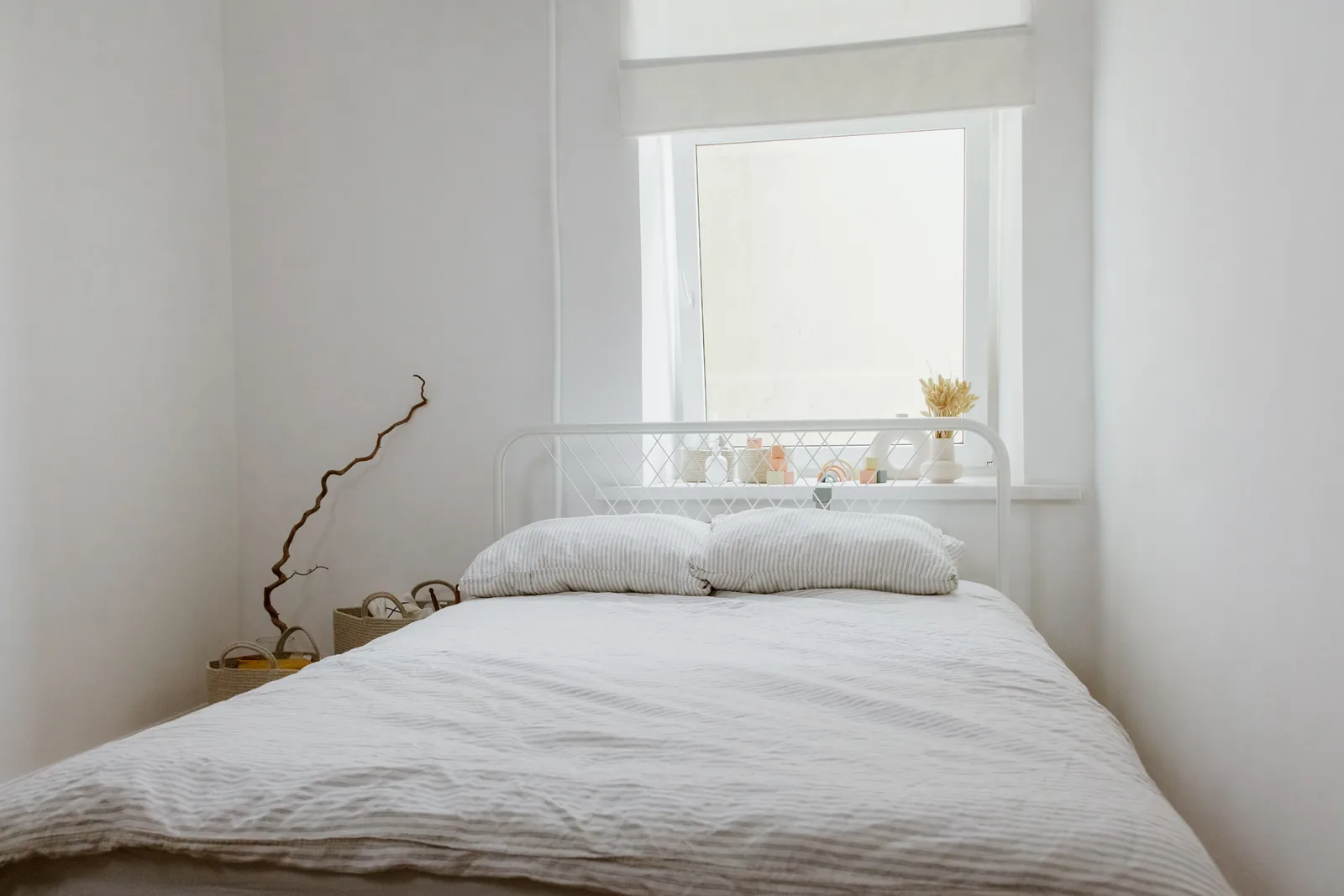 Cama con colcha clara y dos almohadas bajo una ventana en una habitación blanca.